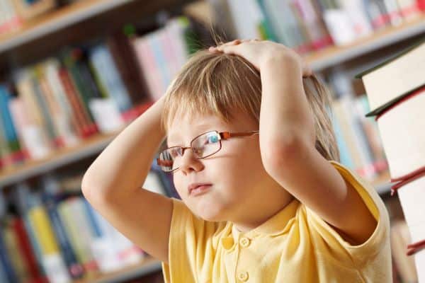 Menino pequeno usando óculos de grau, com as mãos na cabeça, em uma biblioteca escolar.