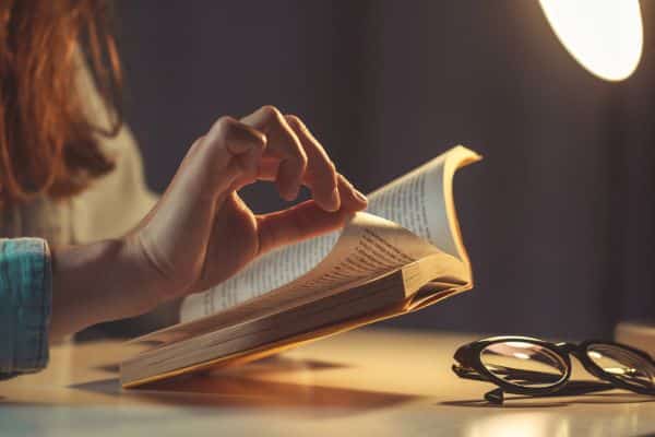 Mão feminina folheando páginas de um livro em local com baixa iluminação. Os óculos estão depositados a sua frente. Conceito de Ler e seus mitos