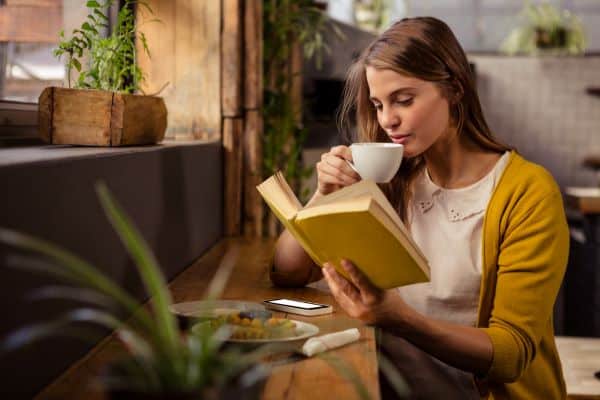 Mitos sobre ler com baixa luminosidade ou em veículo em movimento. Na imagem, jovem mulher lê um livro, frente a uma janela com iluminação natural, enquanto toma um café.