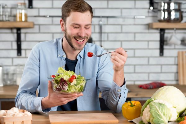 Homem, sentado à mesa em sua cozinha, sorri enquanto se alimenta de forma saudável com uma salada.