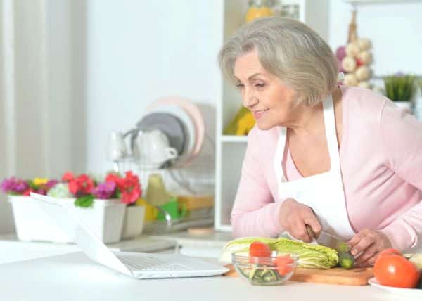 Mulher idosa, com cabelos grisalhos e usando um avental branco sobre uma blusa rosa, sorri enquanto lê um livro de receitas e prepara uma comida saudável