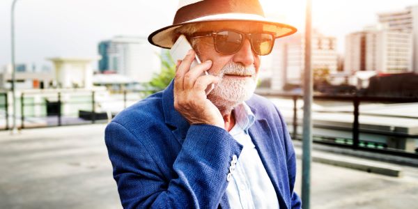 Homem idoso com cabelo e barba grisalhos protege os olhos com chapéu e óculos solar enquanto passeia pelo centro da cidade e conversa pelo telefone celular.