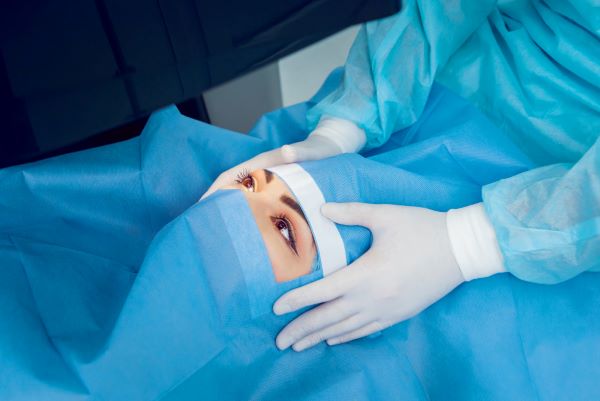 Paciente coberta por tecido azul que deixa apenas à mostra os olhos. Sobre seu rosto, vê-se as mãos do médico, usando luvas brancas, ajeitando o campo visual. 