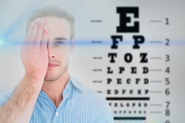 Homem jovem tapa um dos olhos enquanto realiza teste de acuidade visual. ao fundo vê-se a tabela oftalmológica.