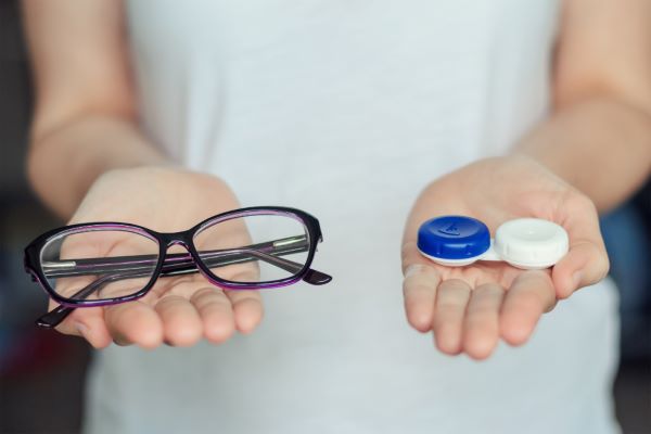 Mulher segura um par de óculos pretos numa mão e, na outra, um estojo de lentes de contato.