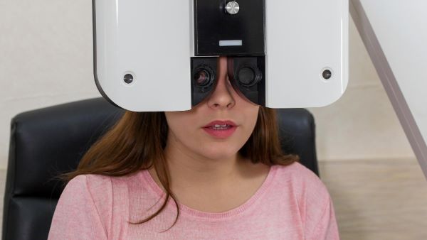 Adolescente, vestindo camiseta rosa, realiza teste por meio de um instrumento de optometria que identifica erros refrativos como a miopia ou a hipermetropia. 