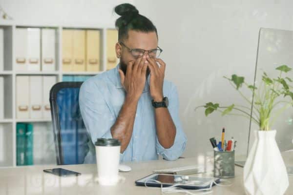 Bonito jovem negro, sentado frente à sua mesa de trabalho em um escritório, coça os olhos levantando os óculos com as pontas dos dedos. 
Coceira intensa é um dos sintomas de doenças que prejudicam a saúde ocular.