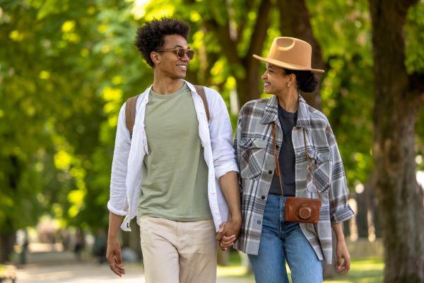 Lindo casal jovem, com roupas primaveris, passeiam de mãos dadas em um parque com muitas árvores. Ambos protegem sua saúde ocular usando óculos de sol - ele- e chapéu - ela.