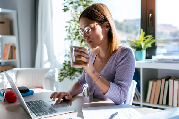 Jovem empresária, com cabelos ruivos e médios, usando óculos e uma blusa lilás, está trabalhando frente a seu computador enquanto segura uma caneca de café.