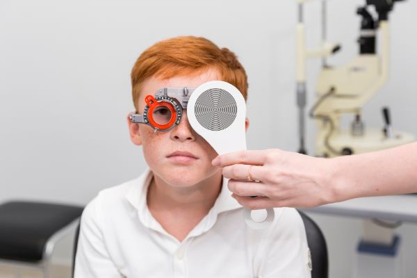 Menino ruivo com sardas no rosto, sério, realiza exames oculares em consultório oftalmológico.