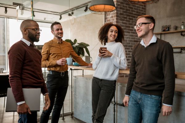 Grupo de pessoas conversando animadamente, na sala do café, durante um intervalo do trabalho. São 3 homens, dos quais 2 usam óculos de grau , e uma mulher.