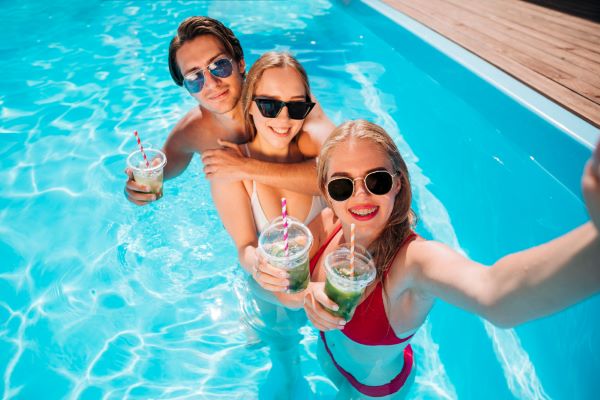 Grupo de amigos, usando óculos de sol e tomando suco, divertem-se fazendo selfies na piscina.