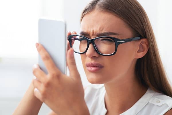 Jovem de cabelos longos e vestindo blusa branca e óculos de grau demonstra dificuldade para enxergar o celular.
Visão embaçada é um dos sintomas das distorções corneanas.