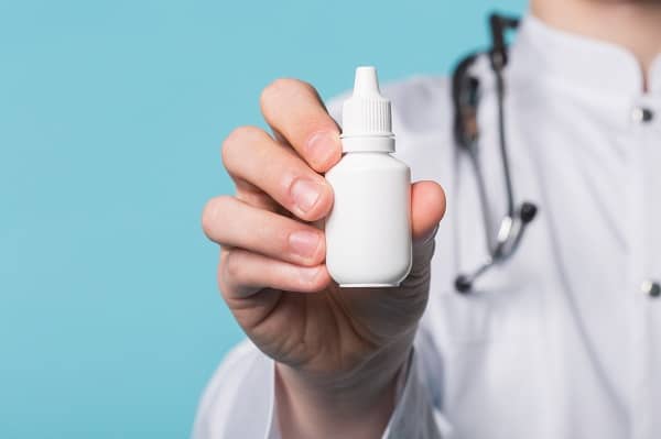 Médico vestindo jaleco branco, e com um estetoscópio pendurado no ombro, segura na mão direita um frasco de colírio.