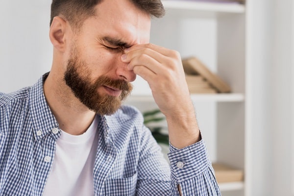 Homem jovem com cabelo, barba e bigode castanhos, vestindo camisa xadrez azul e branco sobre camiseta branca, cerra os olhos em sinal de desconforto ocular.