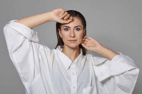 Linda jovem com cabelos molhados e bem maquiada, vestindo uma camisa branca, posa para foto.