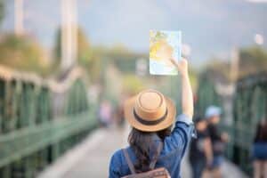 Uma turista, parada em uma ponte longa, levanta com a mão direita um mapa. Ela veste camisa jeans, chapéu e mochila.