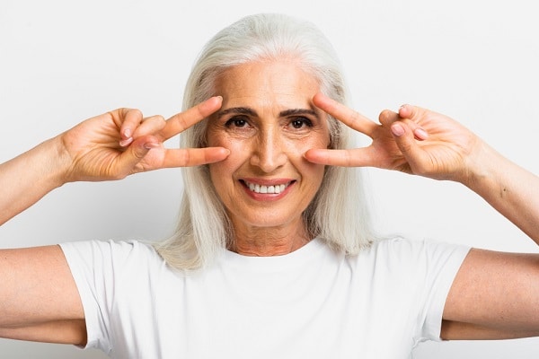 Mulher madura, acima dos 50 anos, sorridente, com cabelos grisalhos, vestindo t-shirt branca, faz sinal de vitória em direção aos olhos.