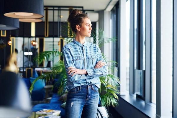 Linda jovem, vestindo calça jeans e camisa listrada azul e branco, com os cabelos presos no alto da cabeça, observa o exterior a partir das janelas do seu escritório.