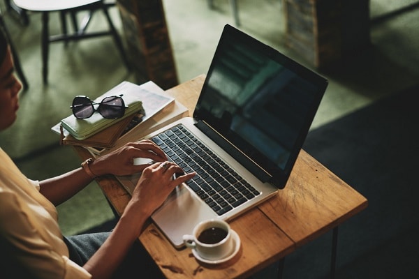 Perfil de uma jovem trabalhando no seu notebook quel se encontra em uma escrivaninha. Também sobre a mesa, uma xícara de cafezinho.