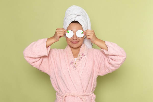 Mulher de roupão rosa sorrindo e cobrindo os olhos com algodão após realização de procedimento. A primavera é o perído ideal para blefaroplastia.